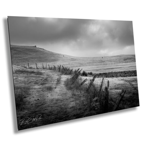 Black and white photo of a weathered fence line winding through rolling California hills under a dramatic cloudy sky.