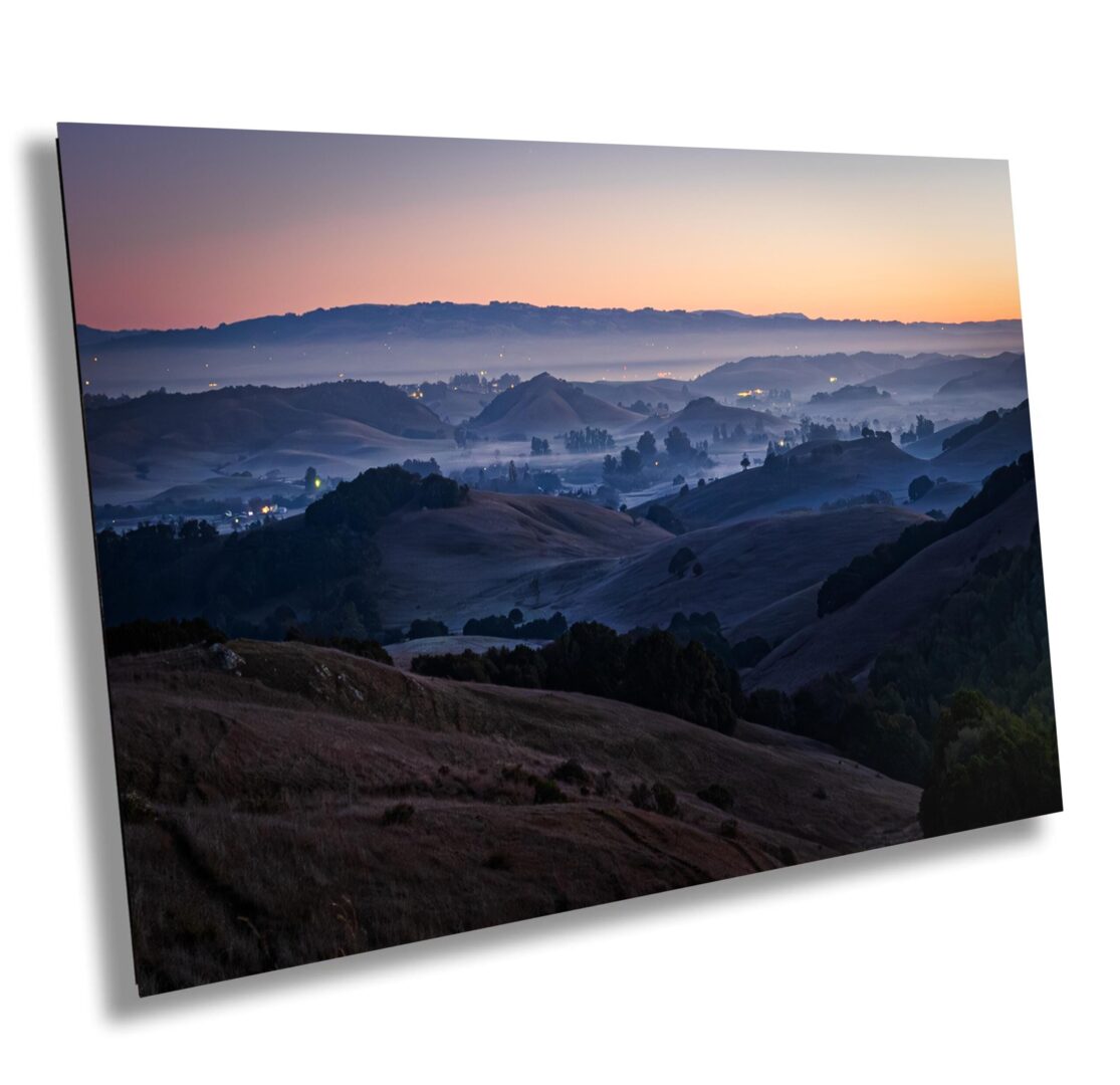 Pre-dawn view of Sonoma County with soft fog drifting through hills and valleys. Blue light, layered ridgelines, and town lights create a peaceful morning scene.
