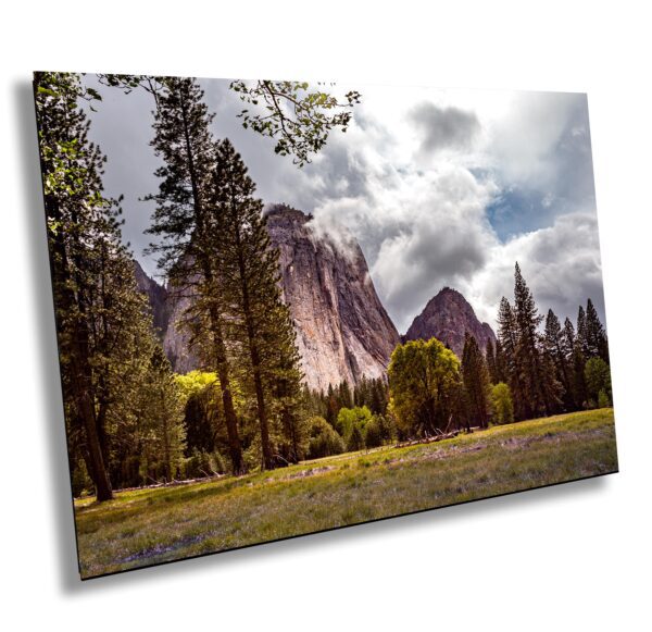 Granite peak in Yosemite National Park framed by pine trees and a grassy meadow under dramatic storm clouds with sunlight breaking through.