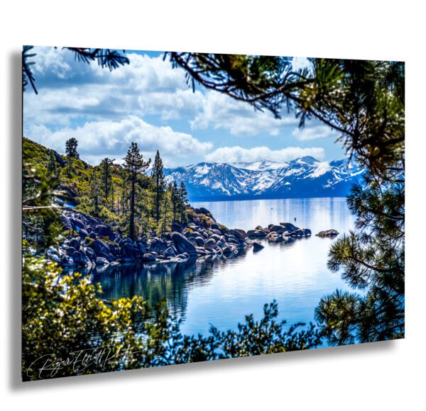 Framed view of Lake Tahoe through pine trees with rocky shoreline, calm reflective water, and snow-covered Sierra Nevada mountains in the distance.