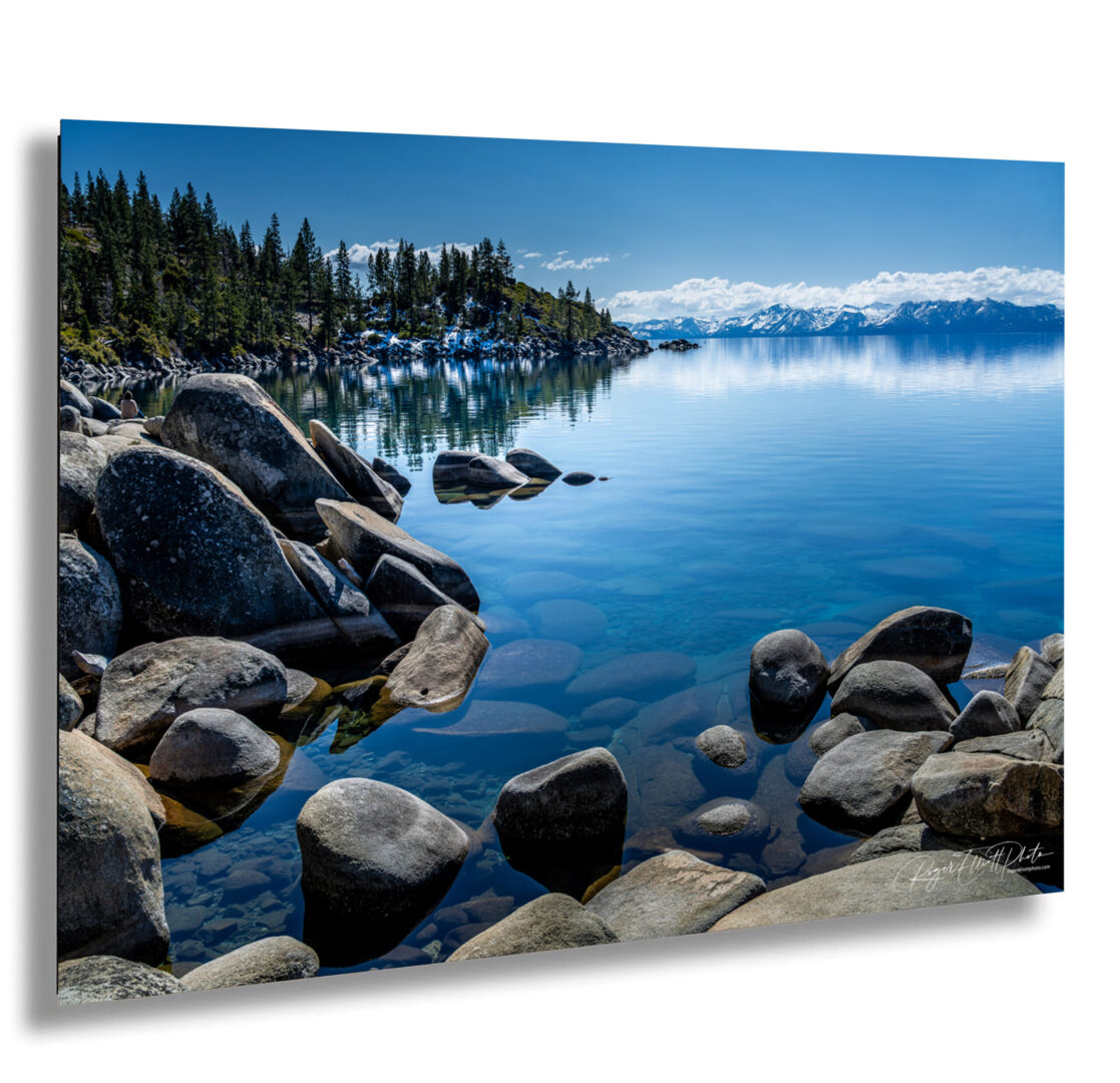 Crystal-clear water and granite boulders along Lake Tahoe’s Nevada shoreline, with pine trees and snow-covered mountains reflected in the calm lake on a sunny day.