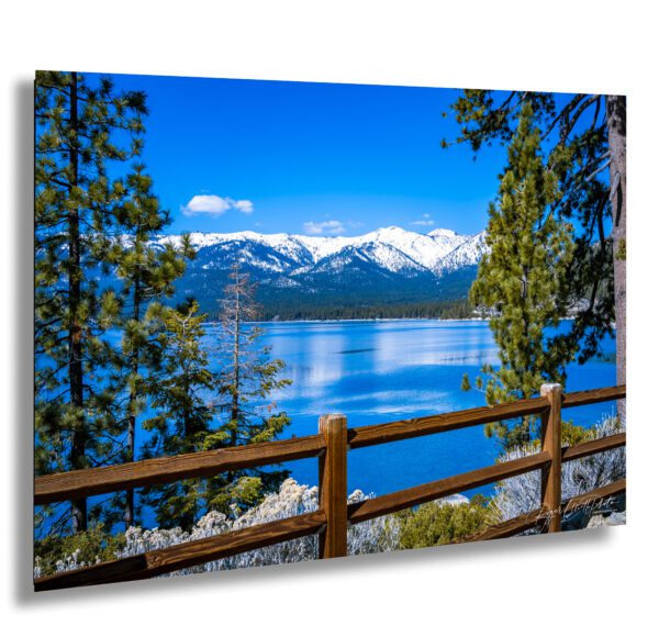 Scenic view of Lake Tahoe with a wooden fence, snow-covered Sierra Nevada mountains, and tall pine trees on a bright blue winter day