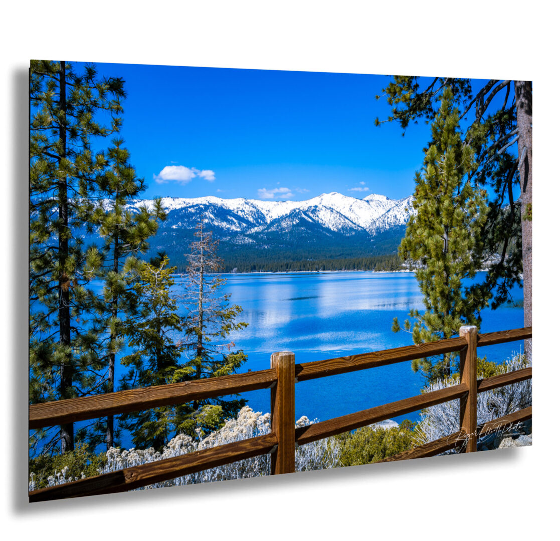 Scenic view of Lake Tahoe with a wooden fence, snow-covered Sierra Nevada mountains, and tall pine trees on a bright blue winter day