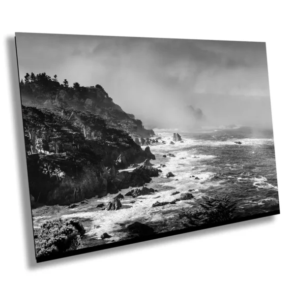 Black and white photo of misty cliffs and stormy ocean waves along the Carmel coast in California, with dramatic sky and rugged shoreline.