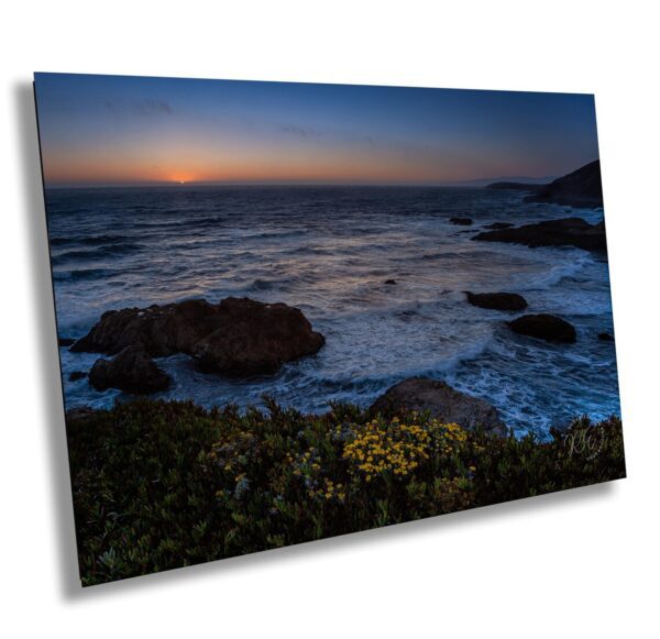 Sunset over the Pacific Ocean with waves crashing on rocks and yellow wildflowers in the foreground along the California coast.